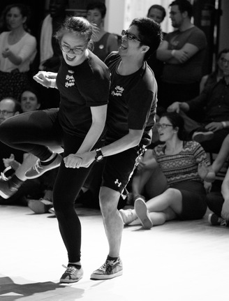 Two dancers in the tandem Charleston, with a crowd in the background behind them. They're one in front of the other, holding hands at their sides, and both standing on their left leg as they lift their right in the air. They have big smiles on their faces, clearly having fun.